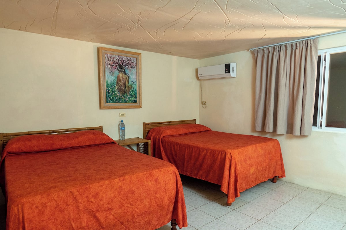 A hotel room with two beds, a painting on the wall, an air conditioning unit, and a window with curtains.