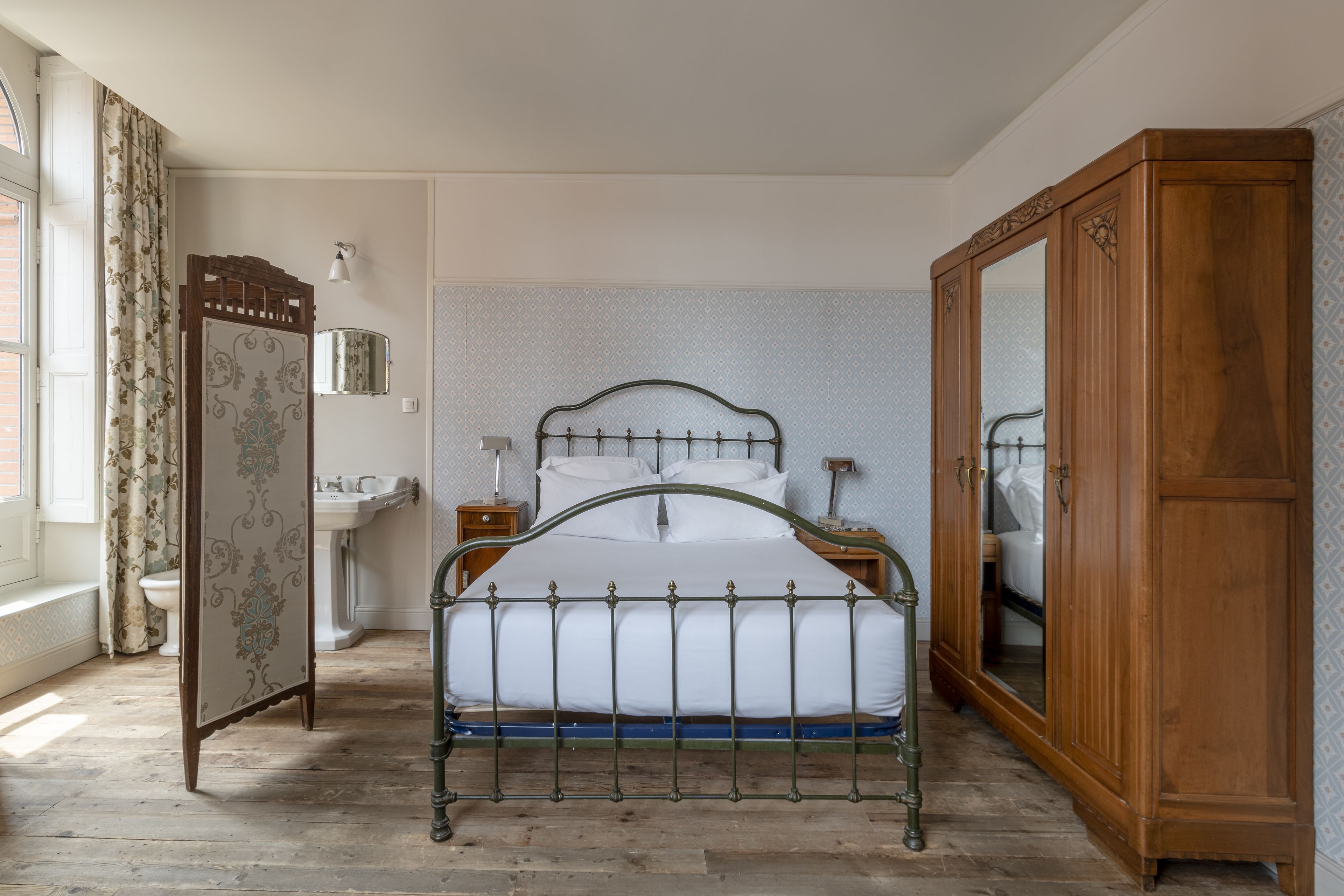 A bedroom with a wooden bed frame, a wardrobe, a bedside table with a lamp, and a bathroom with a sink and mirror.