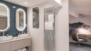 A modern bathroom with two sinks, a large mirror with lights, a towel rack, and a chair with a desk.