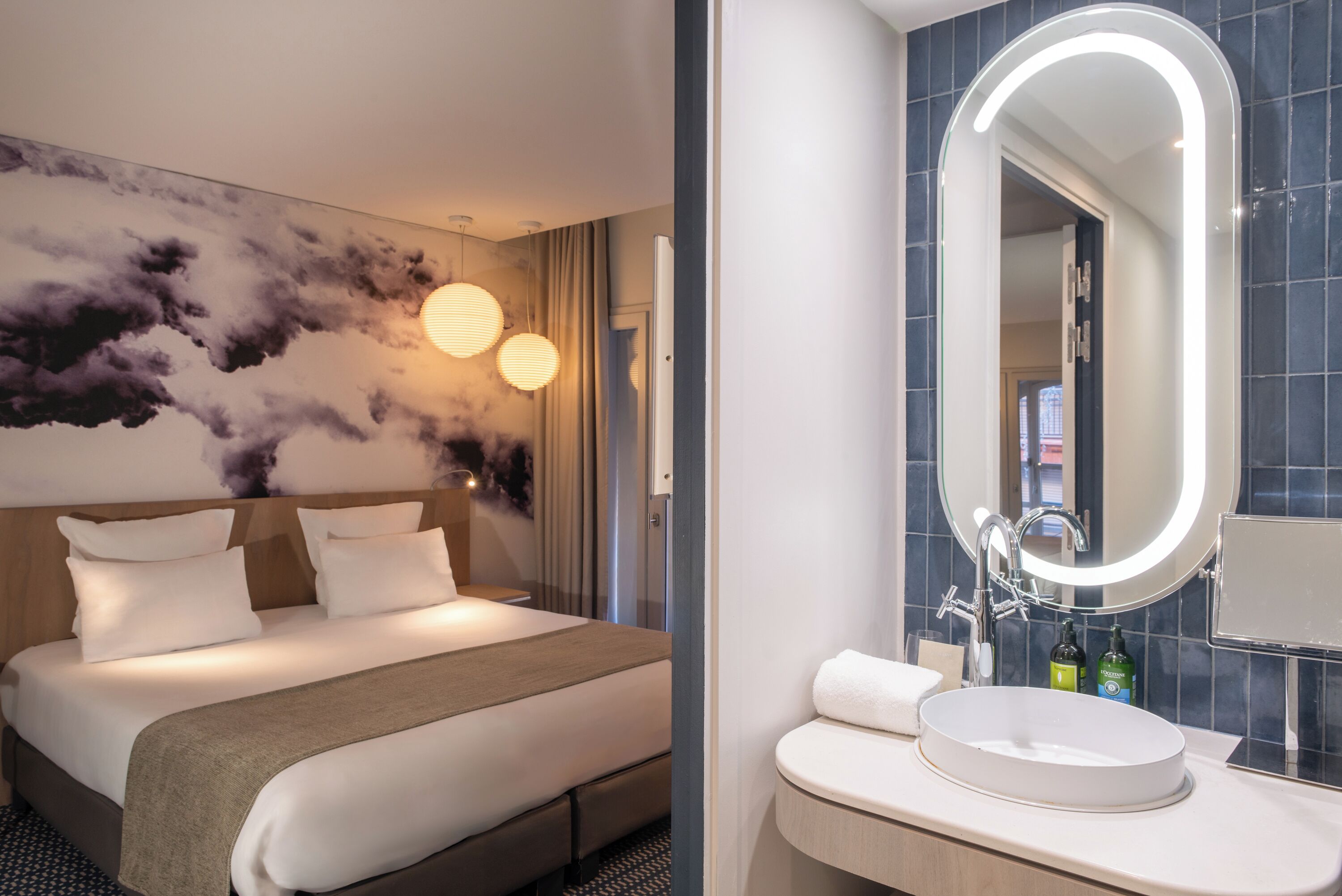 Habitación de hotel con cama, un mural grande en la pared, un espejo redondo y baño con lavamanos y artículos de aseo.