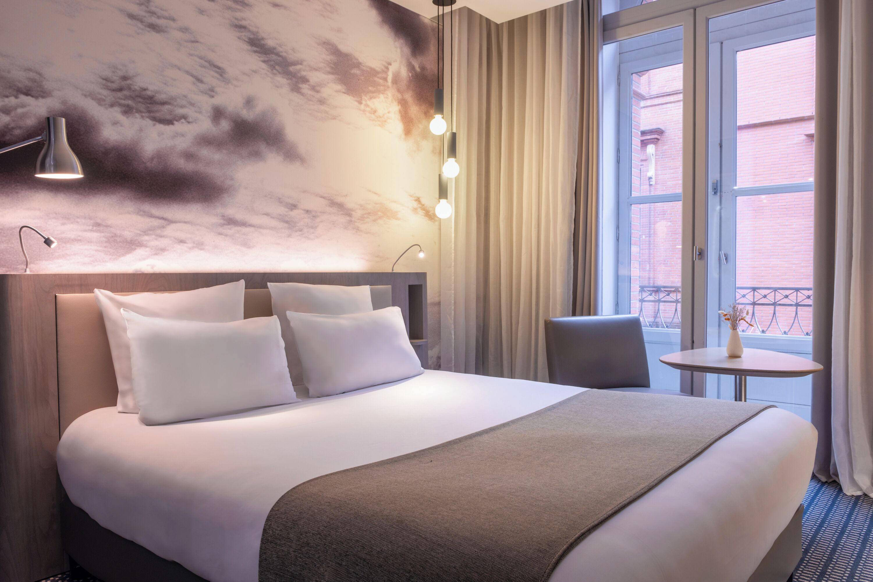 A hotel room with a large bed, a headboard, a bedside table, a chair, a small table with a vase, and a window with curtains.