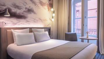 A hotel room with a large bed, a headboard, a bedside table, a chair, a small table with a vase, and a window with curtains.