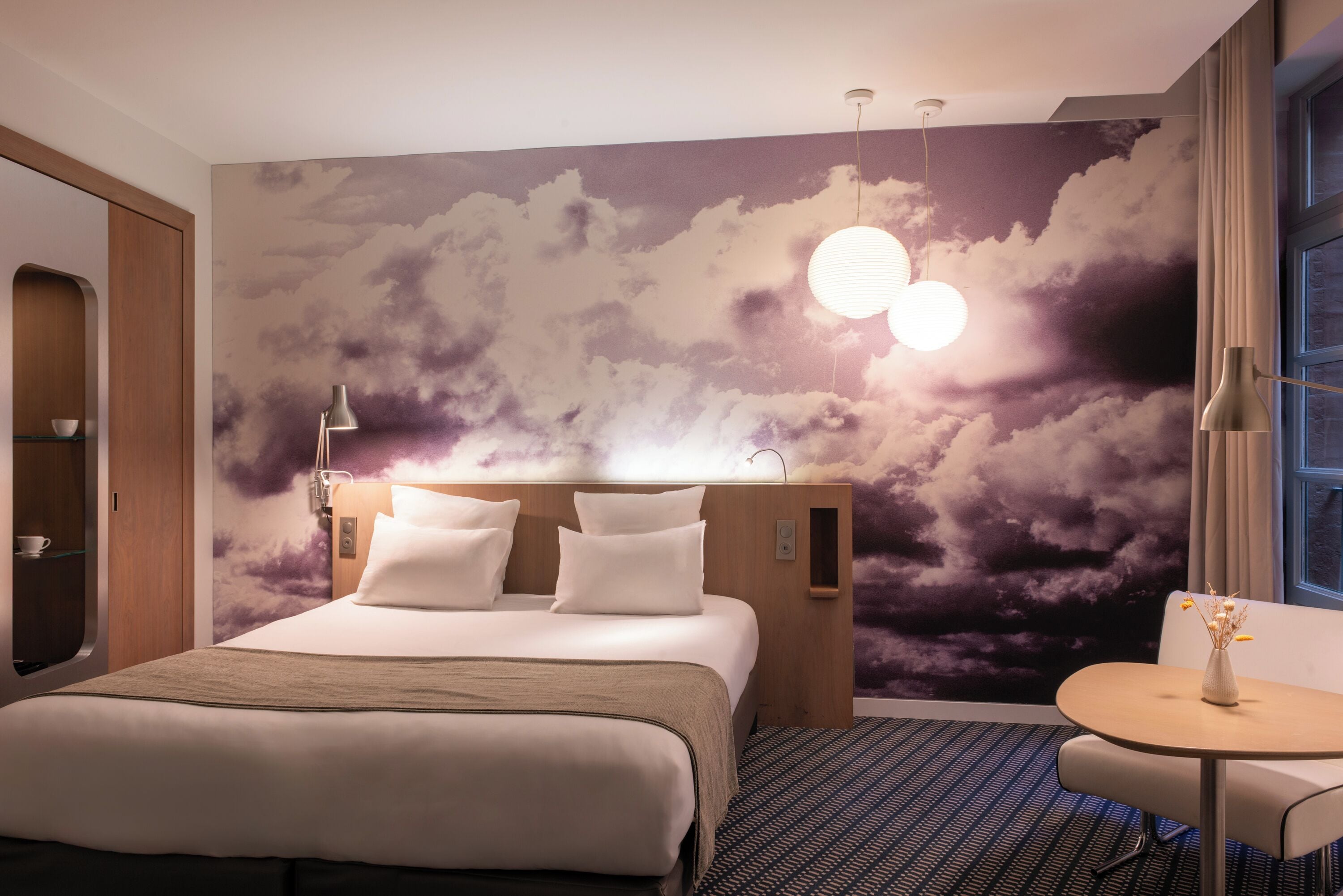 A hotel room with a bed, a bedside table, a chair, a wall mural, and a window.