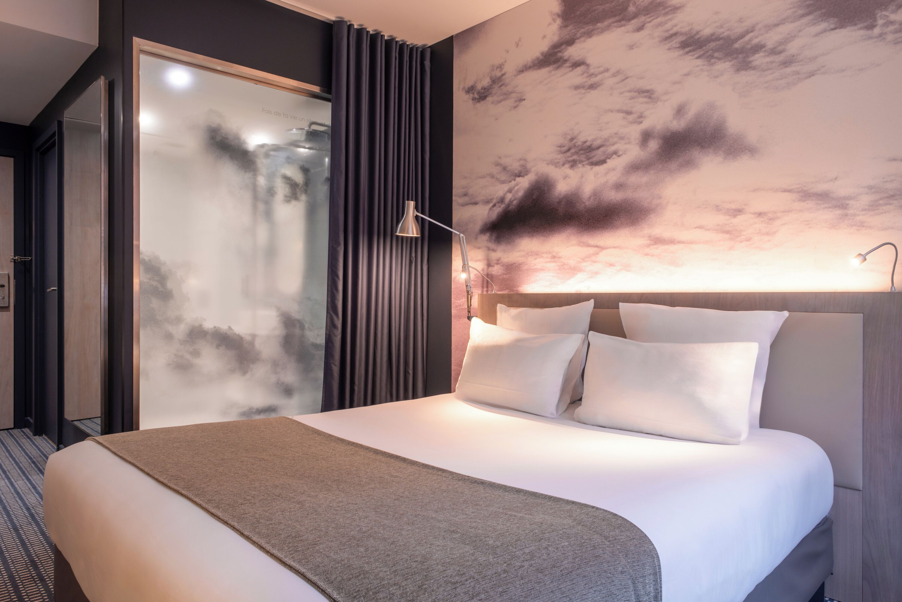 A modern hotel room with a large bed, a glass door leading to a balcony, and a wall mural of clouds.