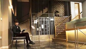 Una persona seduta su una panchina in un'area di accoglienza con una scala e un pannello decorativo.