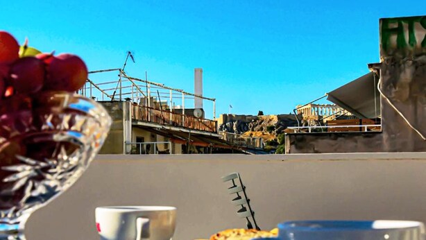 Un tavolo con una ciotola di vetro piena di frutta, due tazze bianche e una fetta di pizza su una terrazza sul tetto, con un cielo azzurro limpido e vista su un edificio storico.