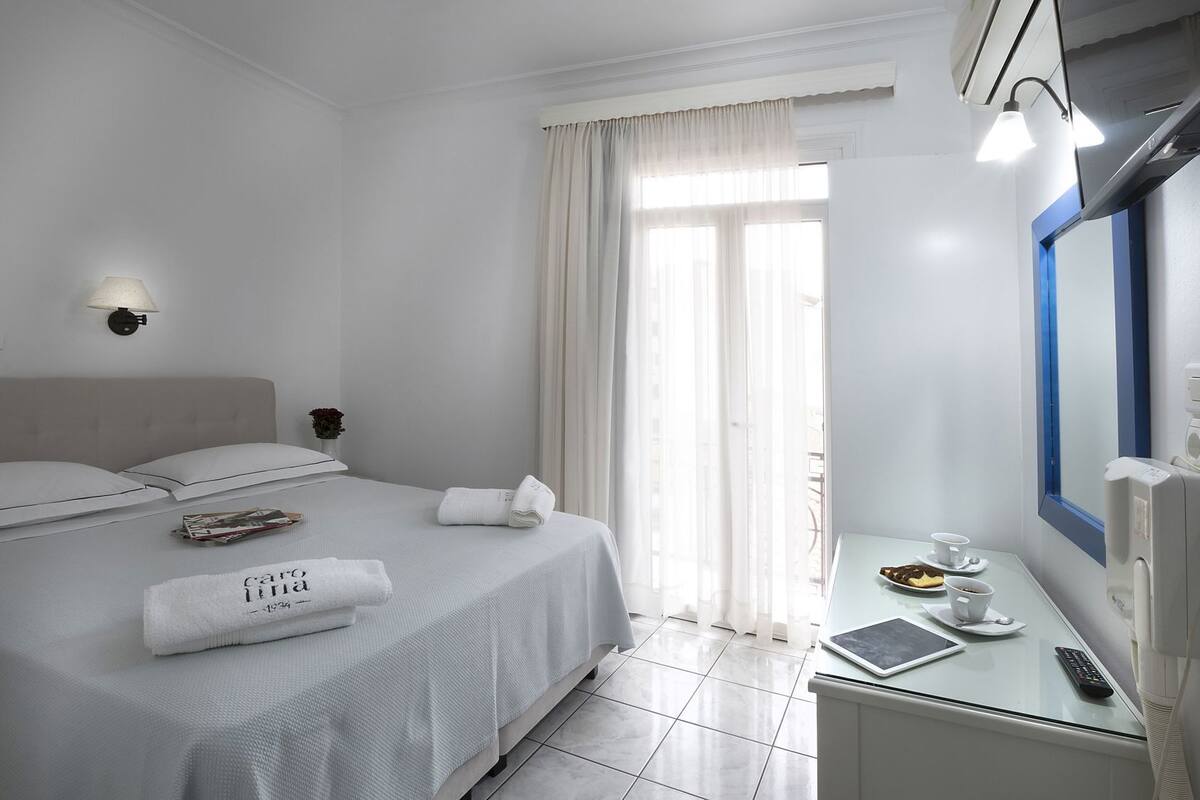 A hotel room with a bed, a bedside table with a remote, a tablet, a cup, and a plate with food.