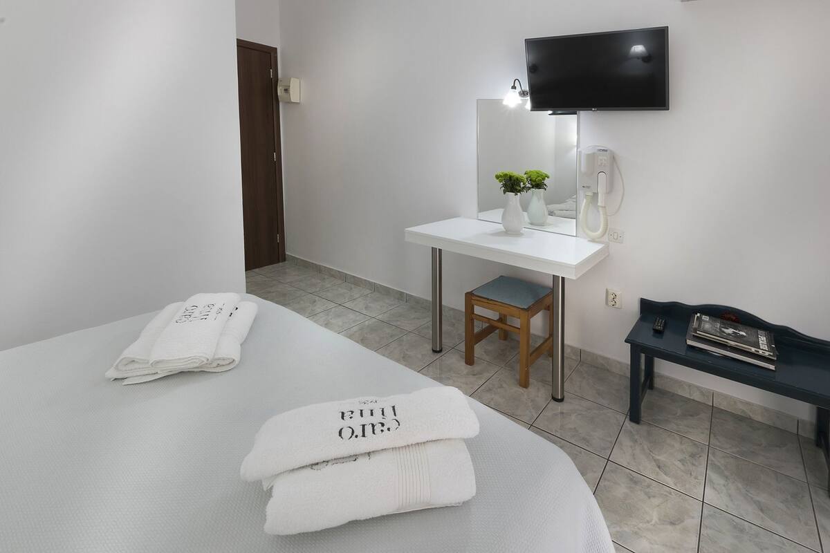 A hotel room with a bed, a desk, a chair, a TV, and a bench.