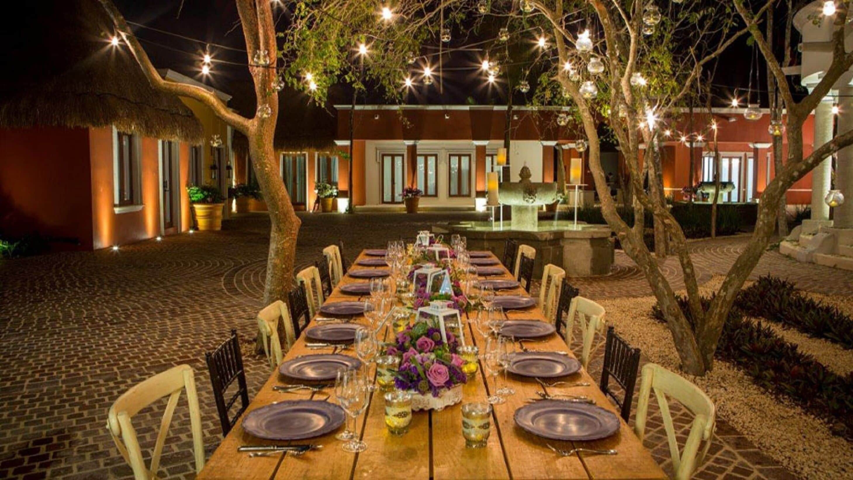 An outdoor dining area with a long wooden table set for a meal, surrounded by chairs.