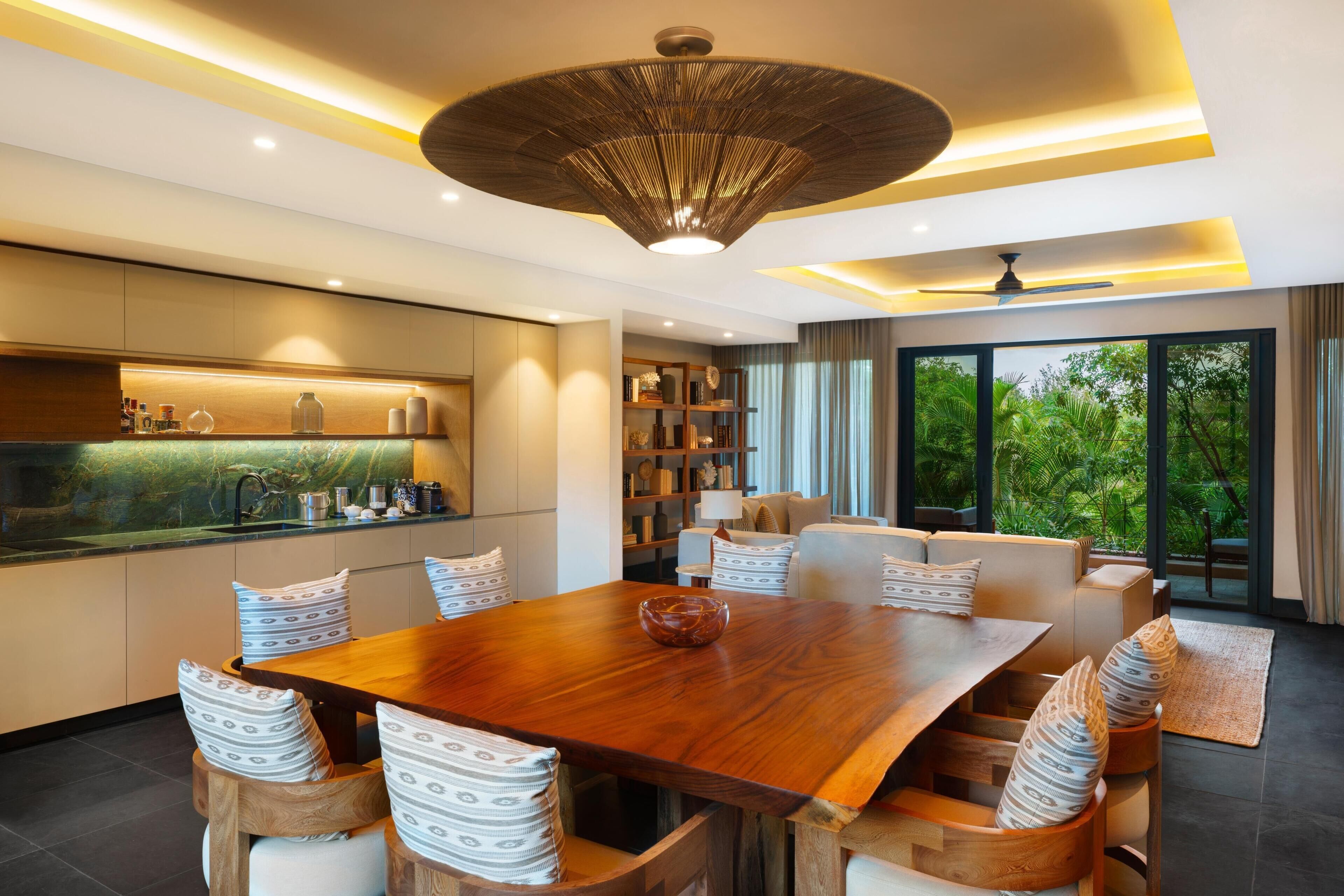 Un comedor moderno con una mesa y sillas de madera, una cocina con salpicadero de mármol y vistas a la vegetación a través de amplios ventanales.