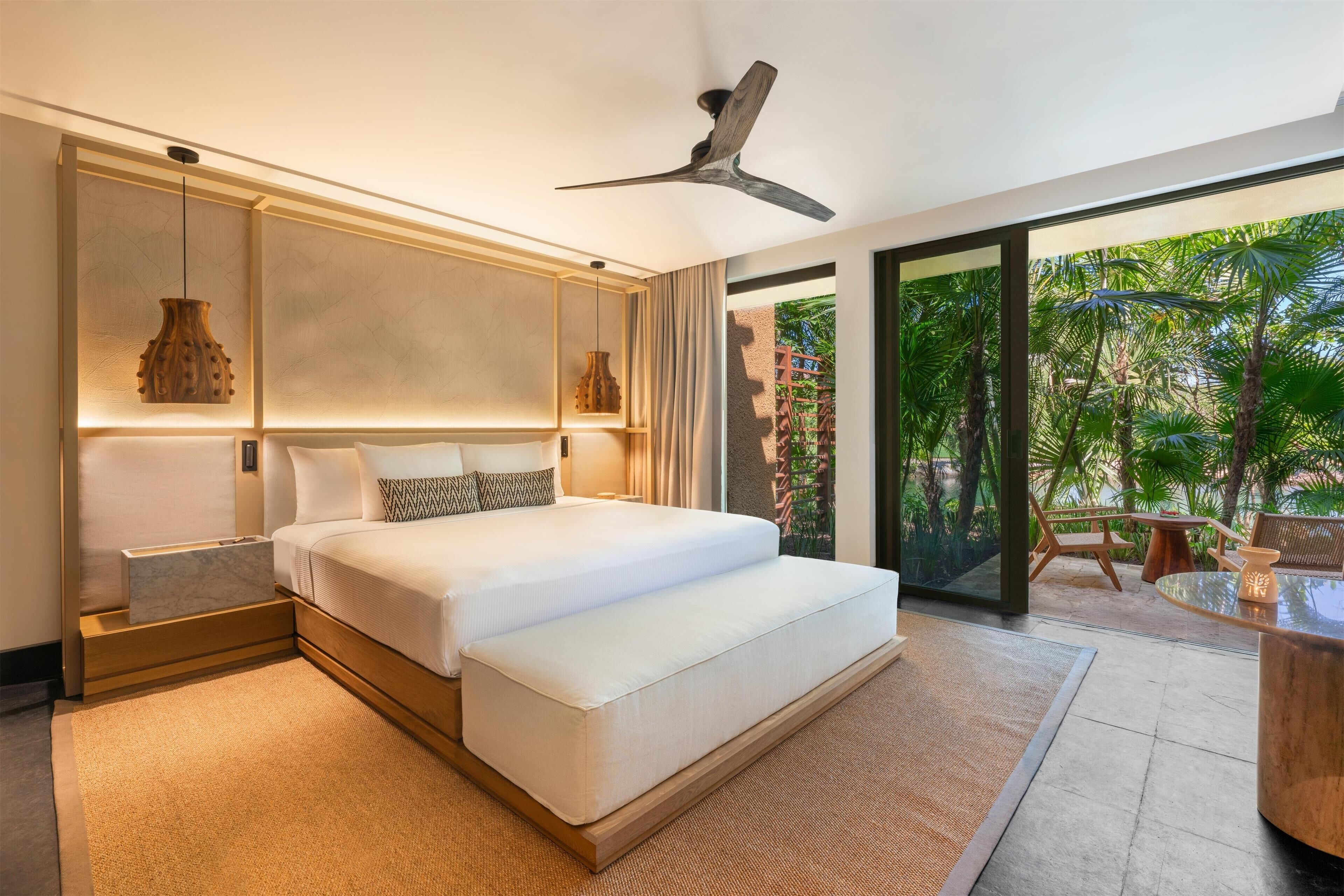 A modern bedroom with a large bed, bedside tables, a ceiling fan, and a view of a tropical garden.