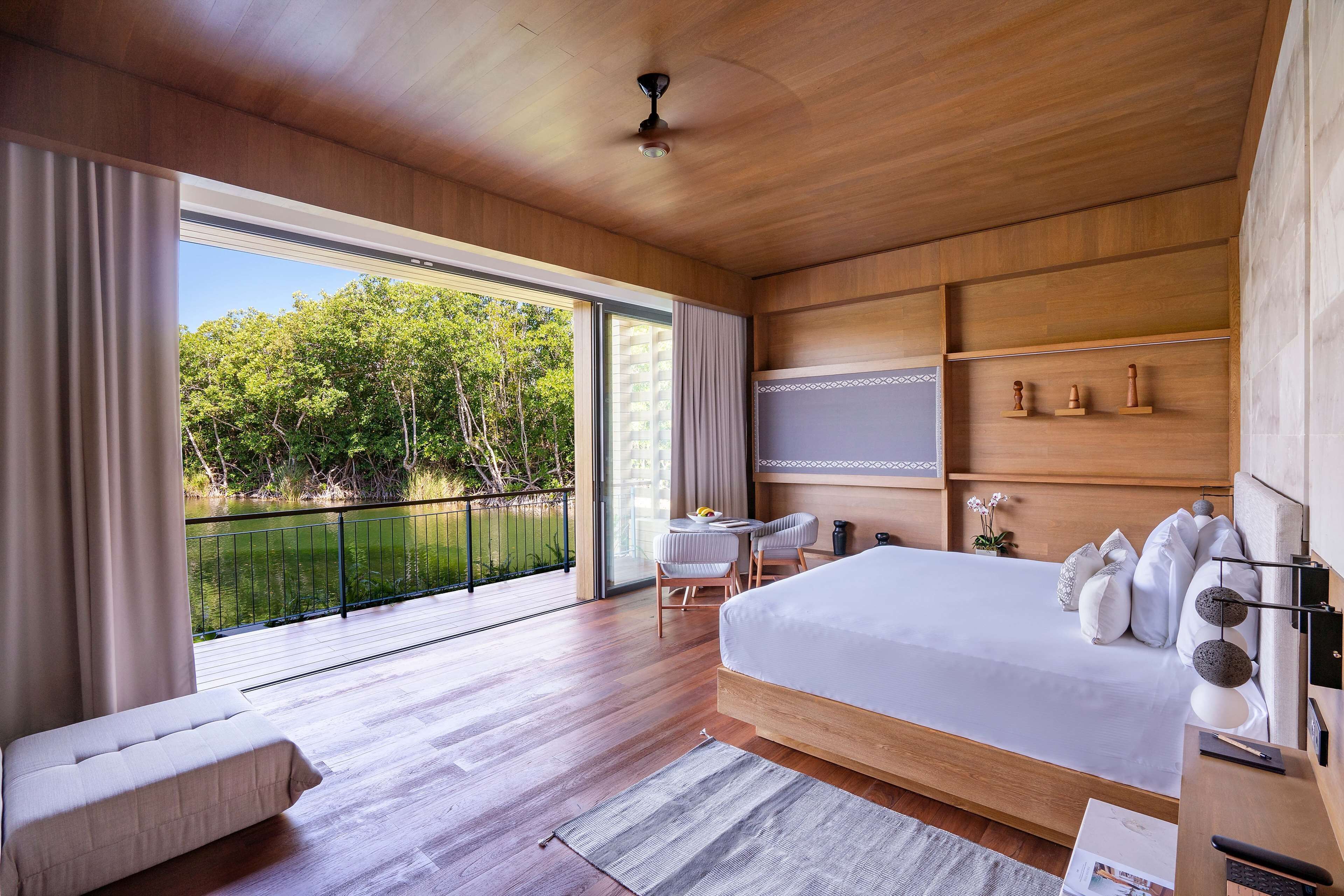 A modern bedroom with a large bed, a balcony with a view of greenery, and a small table with chairs.