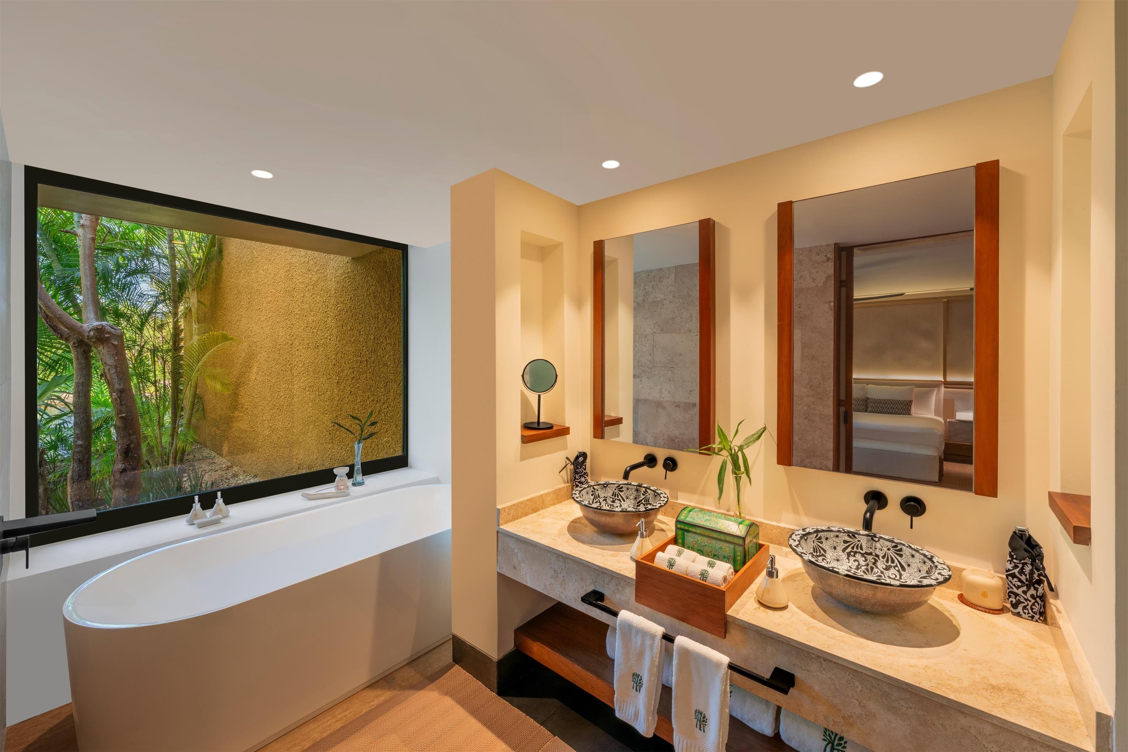 A modern bathroom with a large bathtub, two sinks with decorative bowls, a large mirror, and a window with a view of greenery.