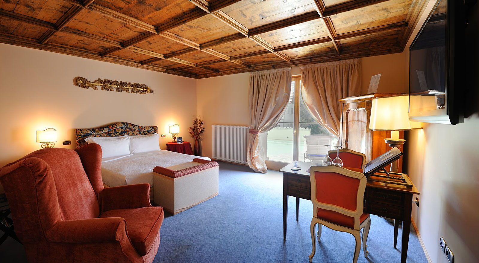 A hotel room with a wooden ceiling, a bed with a floral headboard, a desk with a lamp, and a chair.