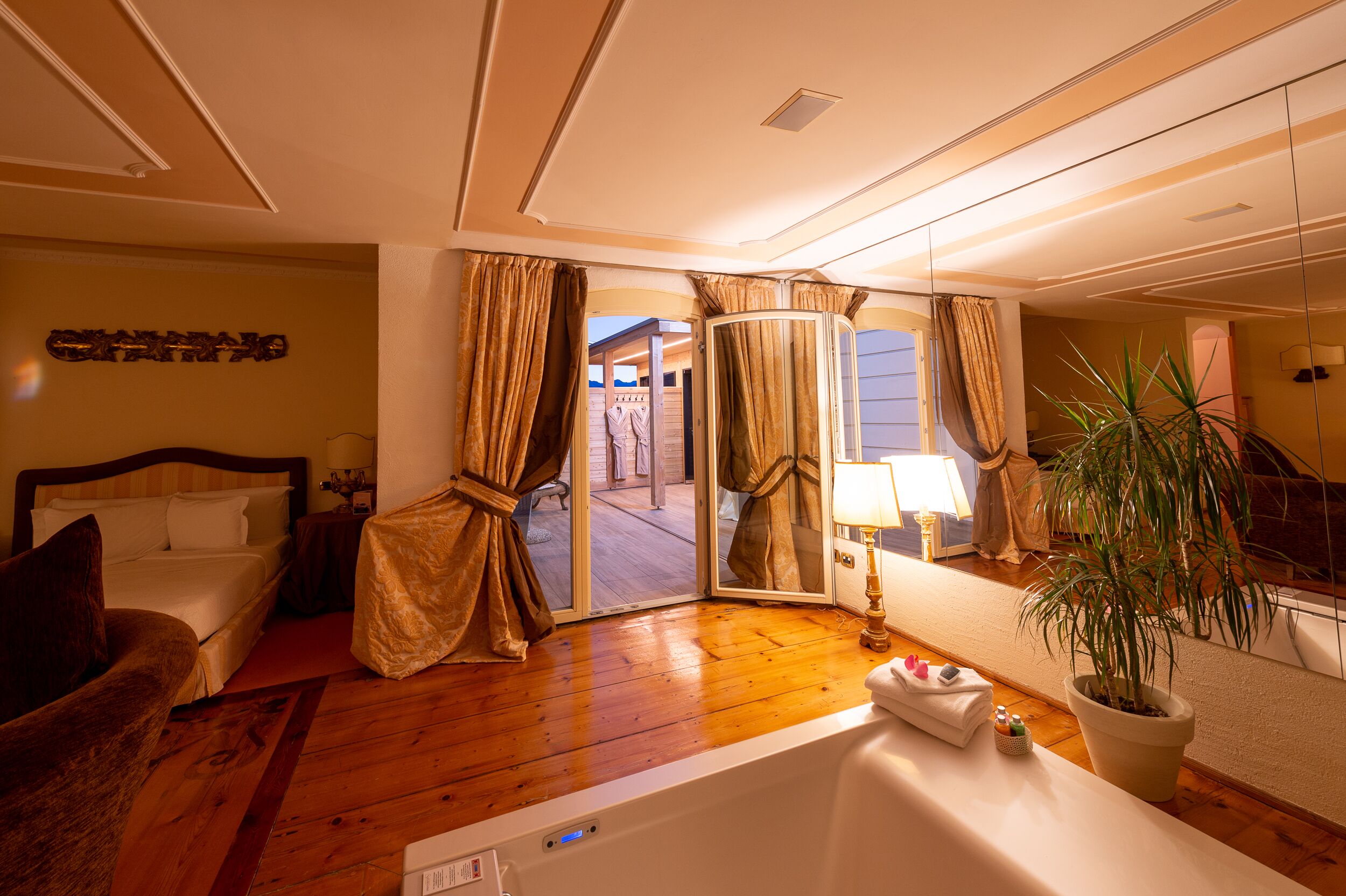 A hotel room with a large bed, a sitting area with a chair, a bathtub, a potted plant, and a mirror reflecting the room.