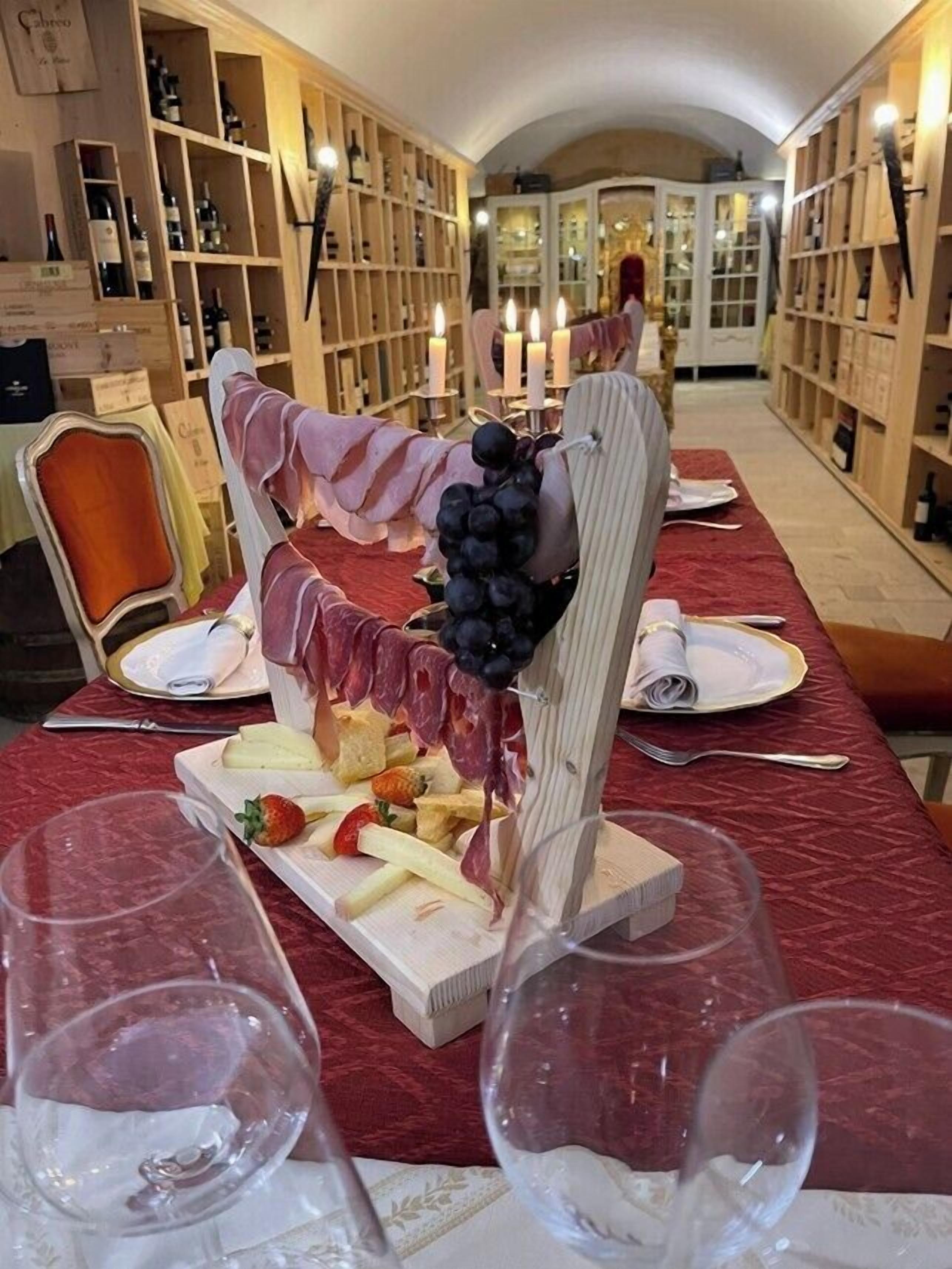 Ein langer Tisch, gedeckt für eine Mahlzeit, mit einer Wurstplatte, Weingläsern und einem Blick auf Weinregale im Hintergrund.