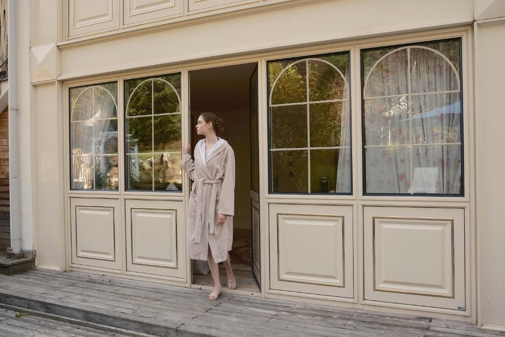 A person in a robe standing at a building entrance with large arched windows and a wooden deck.