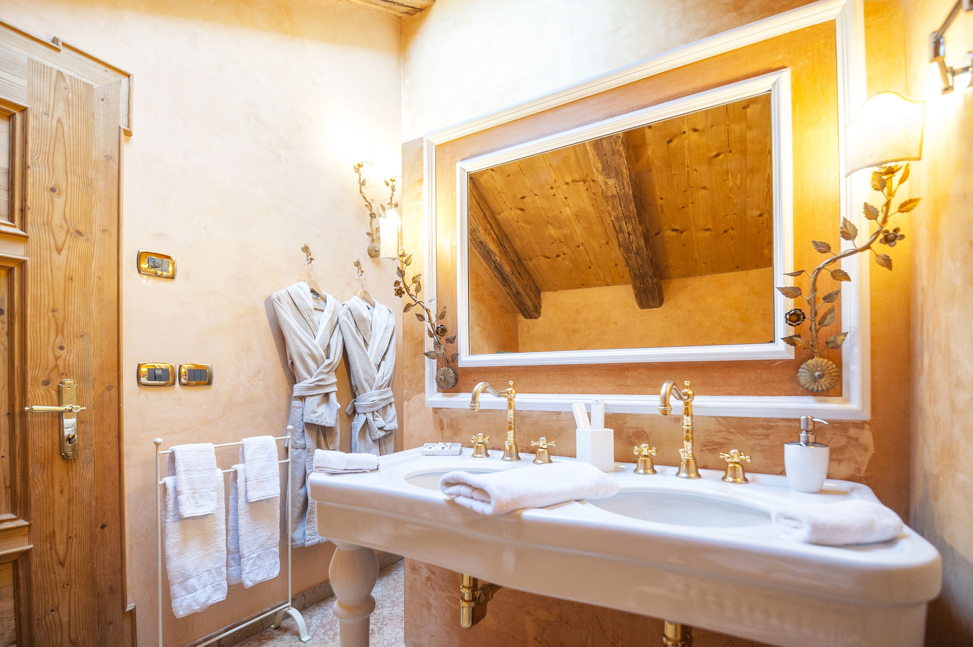 Un baño con techo de madera, un espejo grande y un lavamanos blanco con detalles dorados. Hay toallas en un perchero y una puerta de madera.