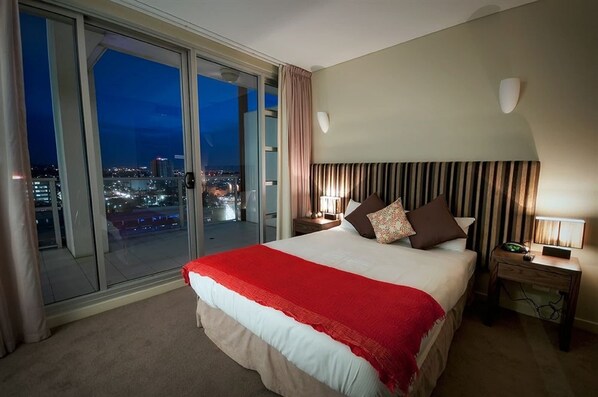 Una habitación de hotel con una cama grande, una mesita de noche, una lámpara y vista a la ciudad de noche.