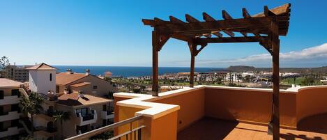 A balcony with a wooden pergola, a tiled floor, and a view of the sea and buildings.