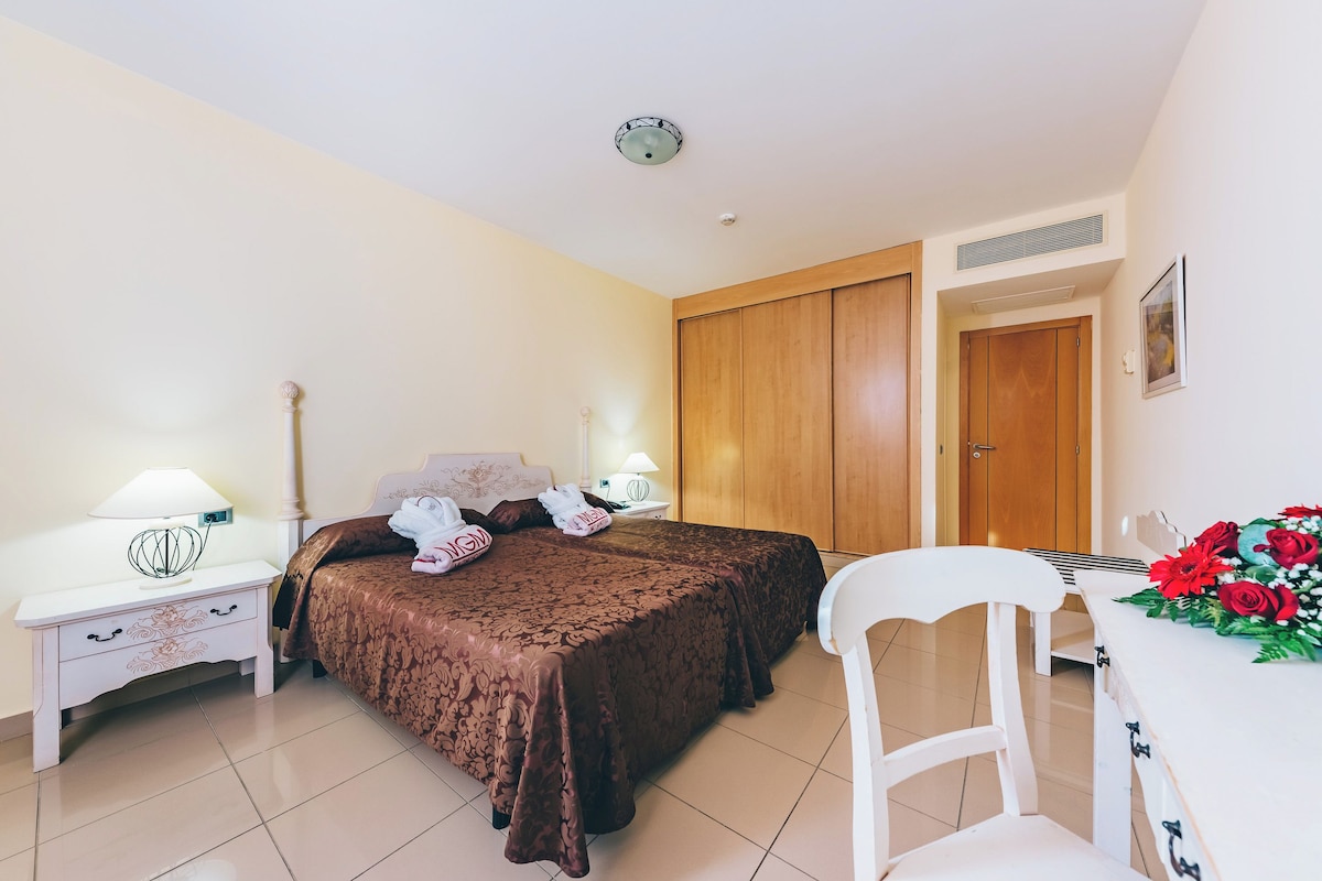 Un dormitorio con cama, mesitas de noche, una silla y un escritorio con un arreglo floral.