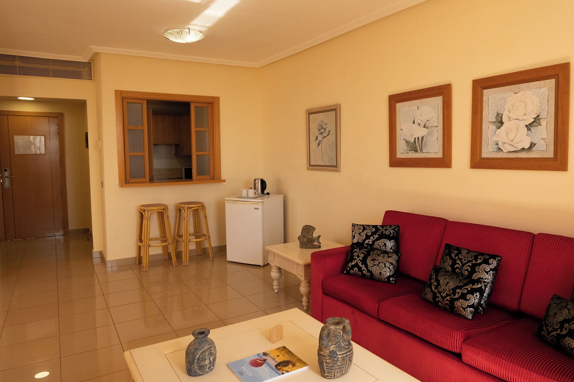 A living room with a red sofa, two stools, a white coffee table, and a small white refrigerator.