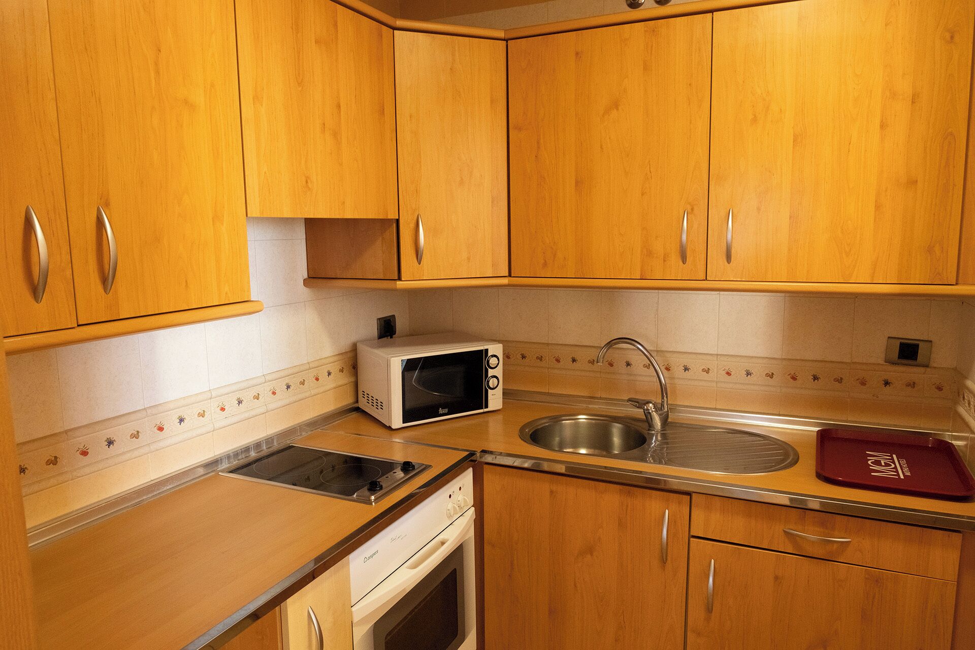 Une cuisine compacte avec des meubles en bois, un four à micro-ondes encastré, un évier en acier inoxydable et un plateau rouge.