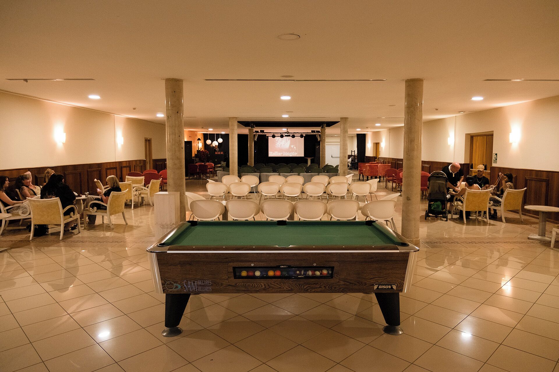 A pool table in a room with chairs arranged around it, a stage with a screen, and a few people seated.