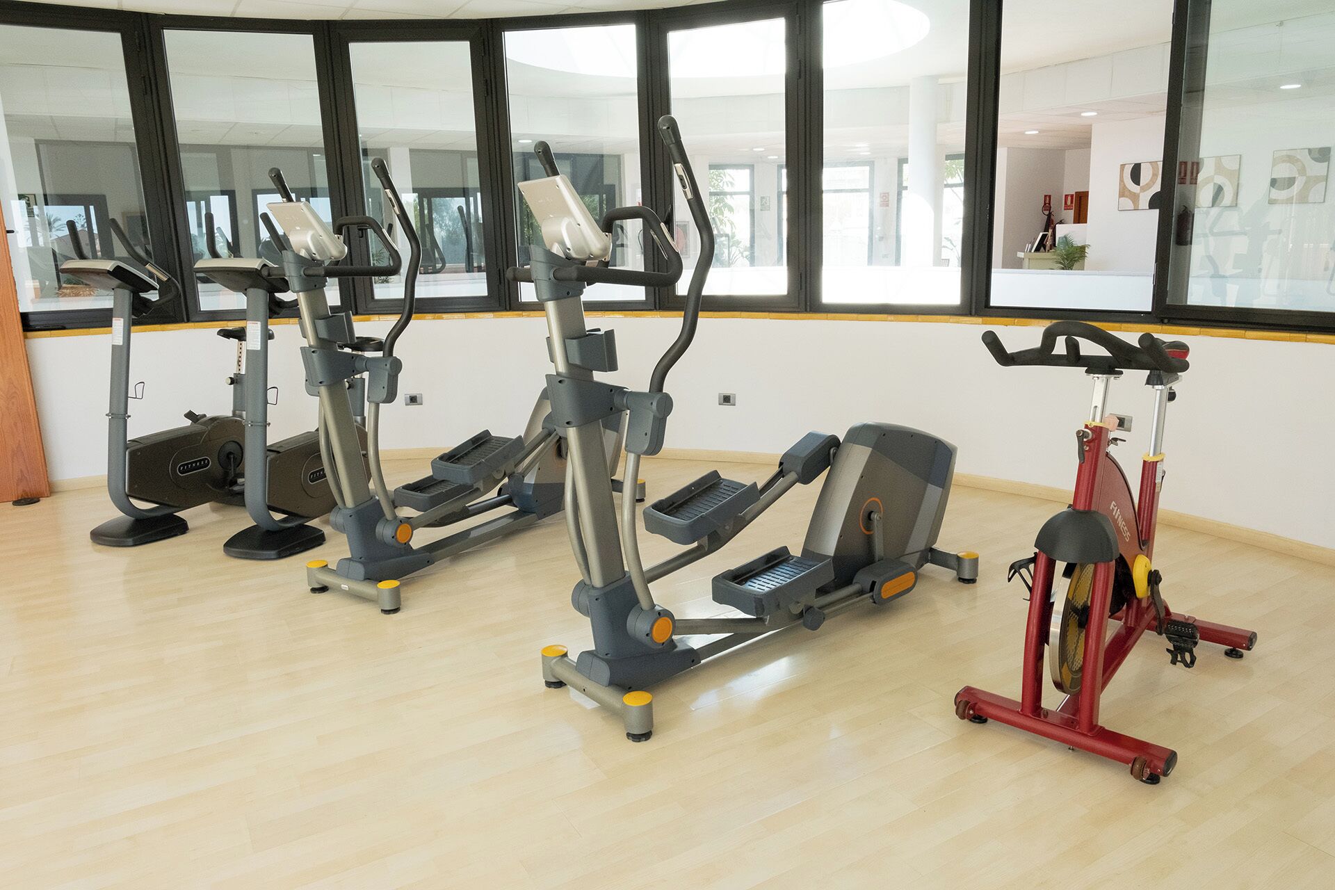 Ein Fitnessstudio mit verschiedenen Trainingsgeräten, großen Fenstern und Holzboden.