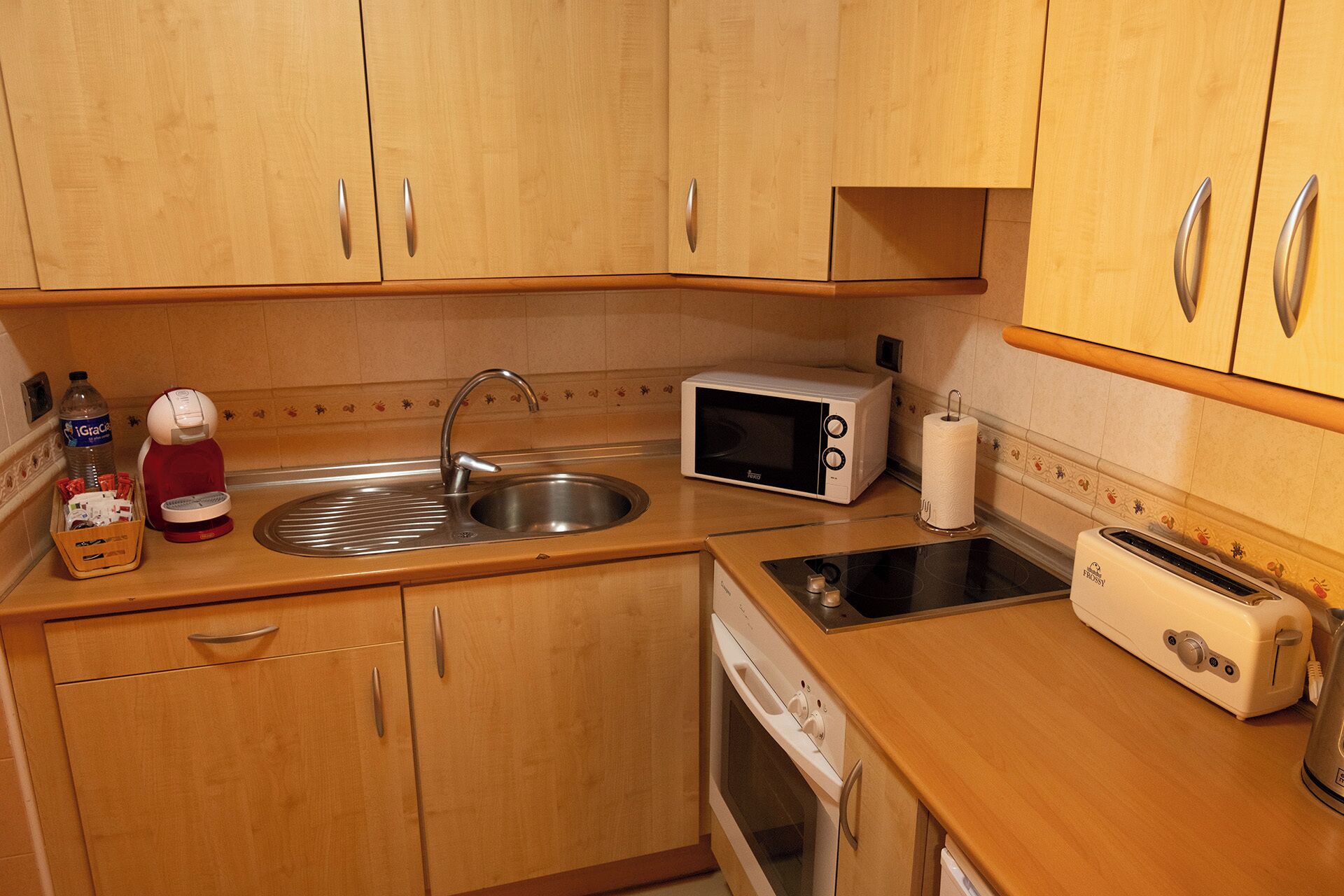 A compact kitchen with wooden cabinets, a stainless steel sink, a microwave, a toaster, and a dish drying rack.