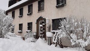 A snow-covered building with multiple windows and a door, surrounded by snow-laden trees and bushes. - Hotel Das Landhaus (Hoechenschwand)