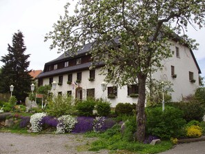 Un bâtiment blanc doté d’un toit en pente, de nombreuses fenêtres et d’un jardin où poussent diverses plantes et fleurs.