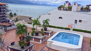 Zona de piscina en la azotea con vista al mar, palmeras y un edificio con cúpulas azules.