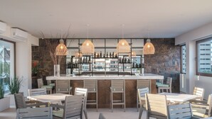Zona de bar moderna con pared de acento de piedra, múltiples lámparas colgantes y diversas opciones de asientos, incluyendo taburetes y mesas.