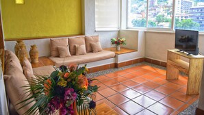 Una sala de estar con un sofá, una mesa de centro, un televisor sobre un mueble de madera y una ventana con vistas a edificios.