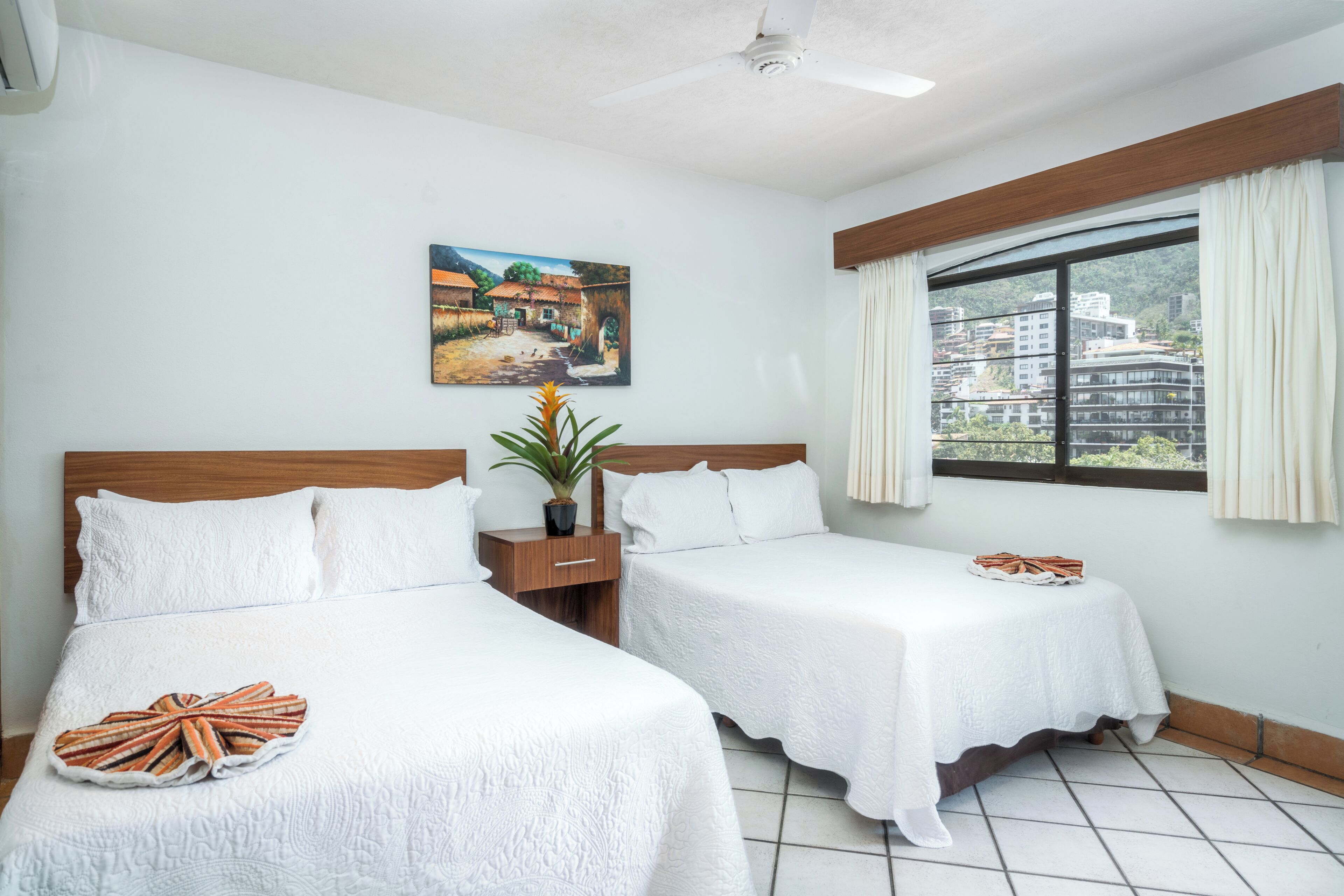A hotel room with two beds, a window with curtains, a ceiling fan, a painting on the wall, and a plant on a nightstand.