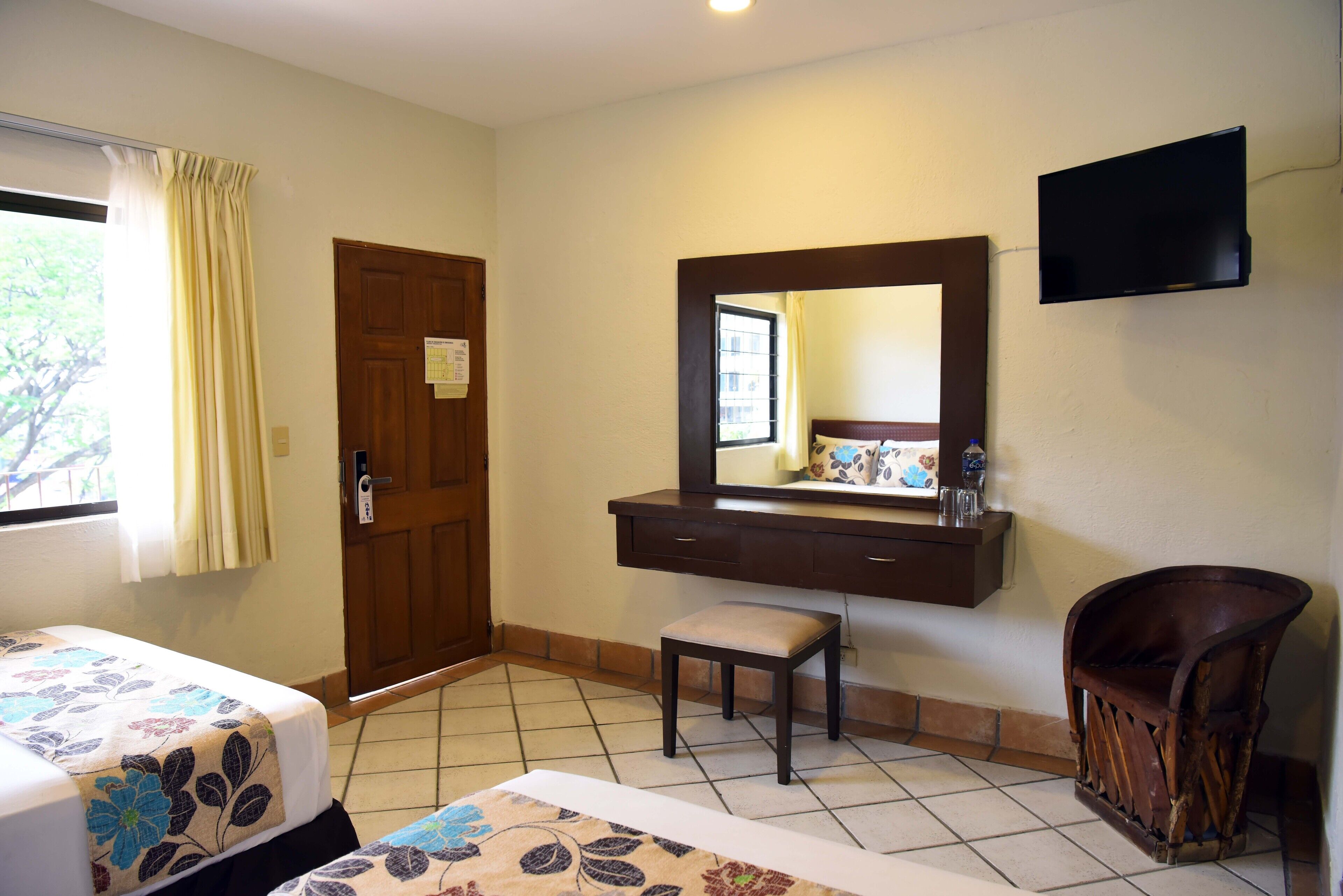 A hotel room with two beds, a wooden door, a small stool, a television mounted on the wall, and a window with curtains.