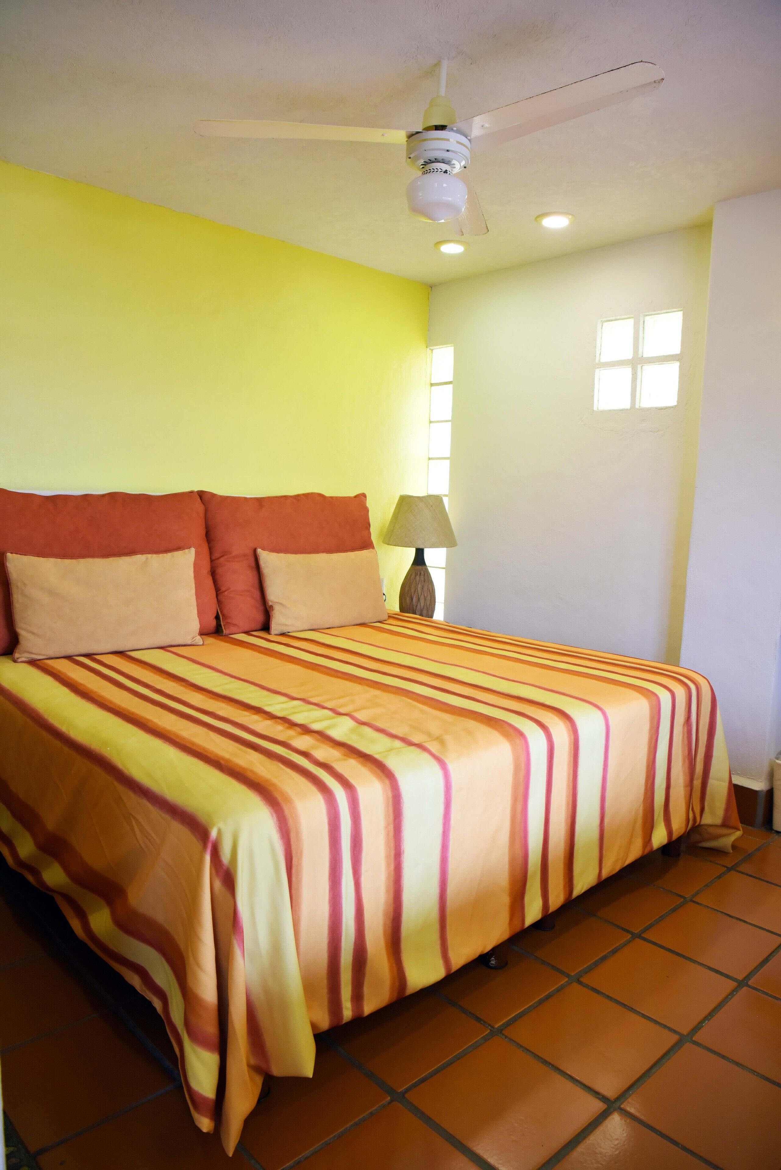 Una cama con una colcha a rayas, un ventilador de techo y una mesita de noche con lámpara.
