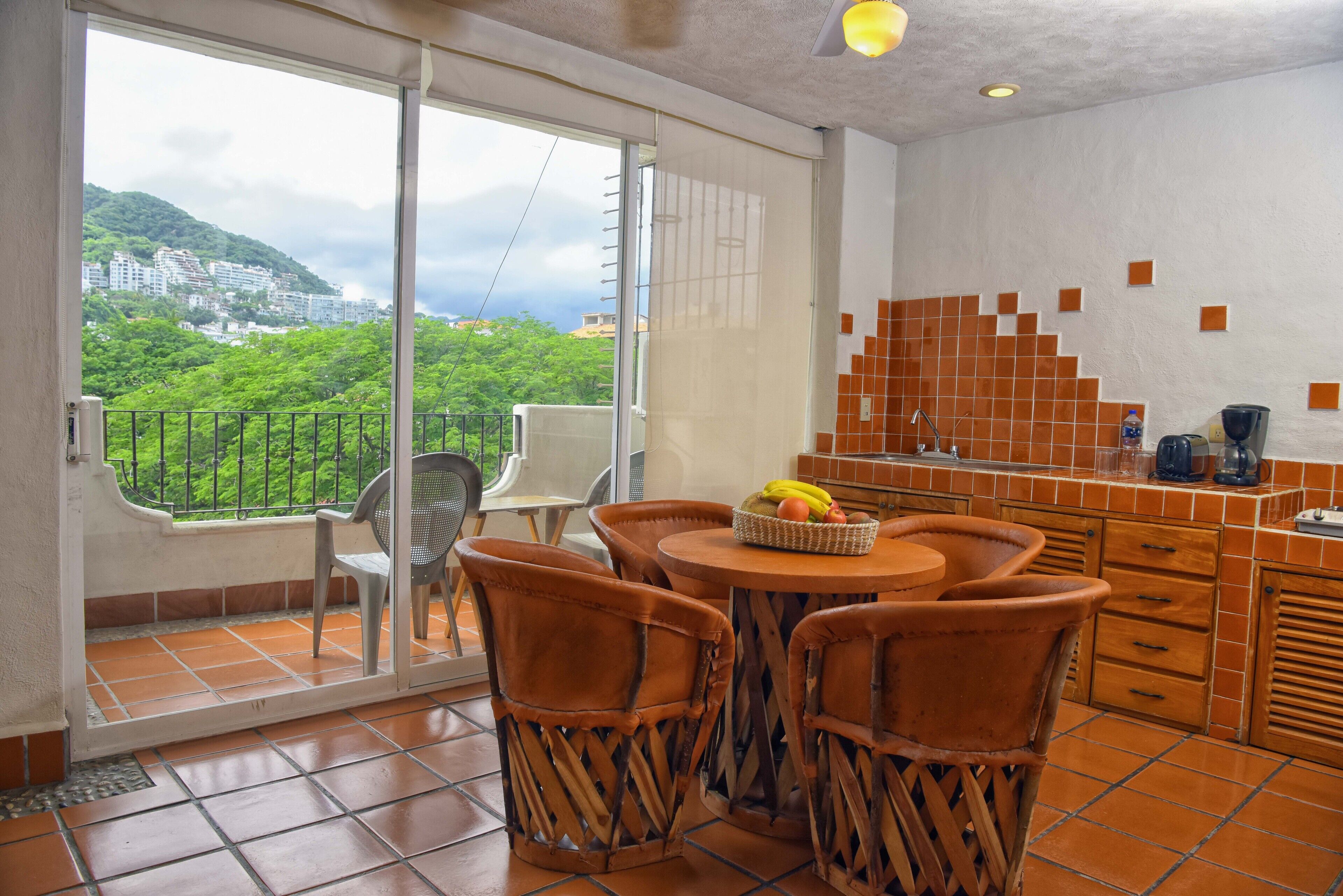 Een balkon met uitzicht op een groene heuvel en gebouwen, een eethoek met rieten stoelen en een tafel, en een keuken met oranje tegels en een koffiezetapparaat.