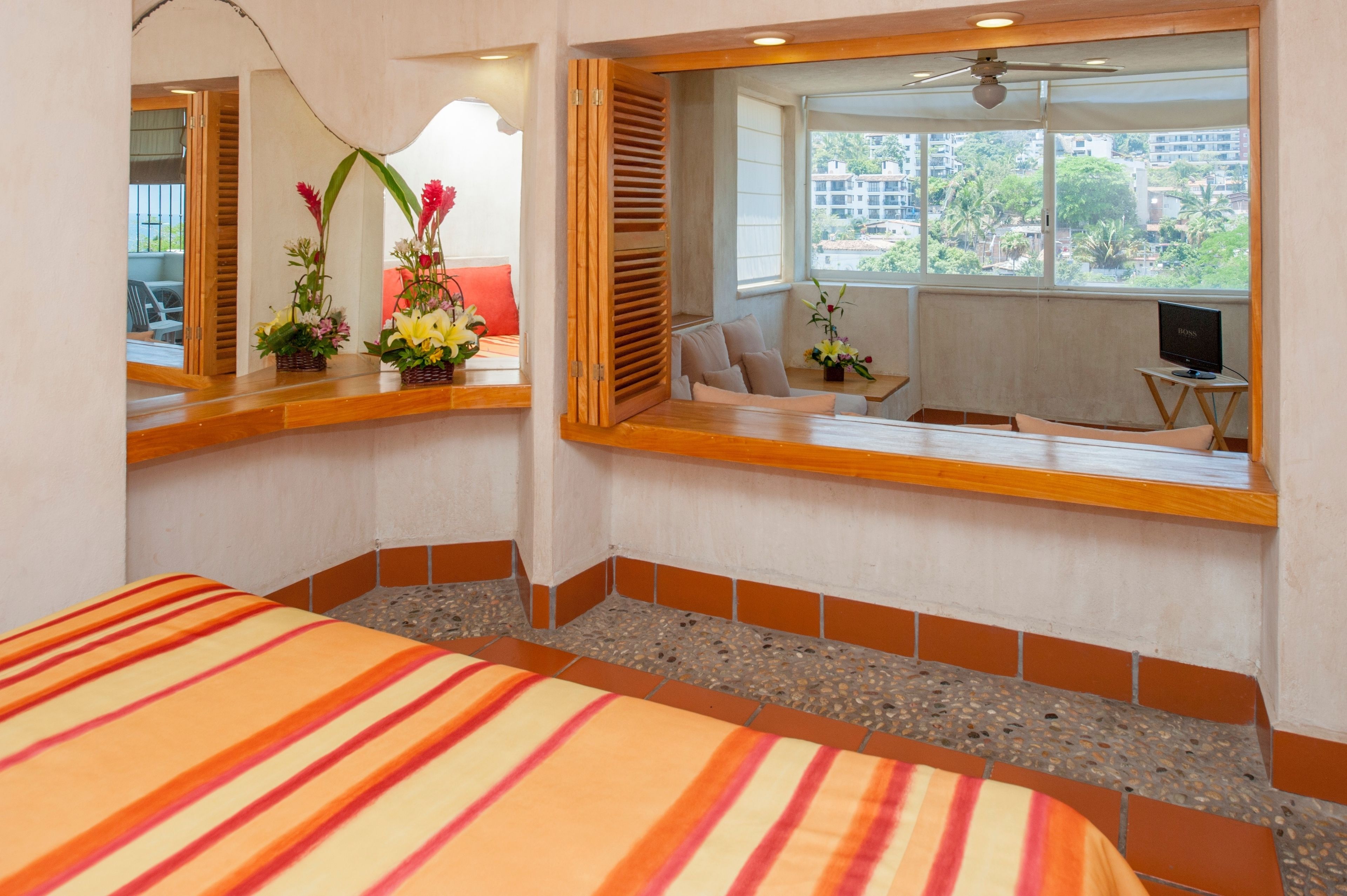 Une chambre d’hôtel avec un lit, une petite table sur laquelle se trouve un téléviseur, une fenêtre donnant sur des immeubles et des arbres, et une plante décorative posée sur le rebord de la fenêtre.