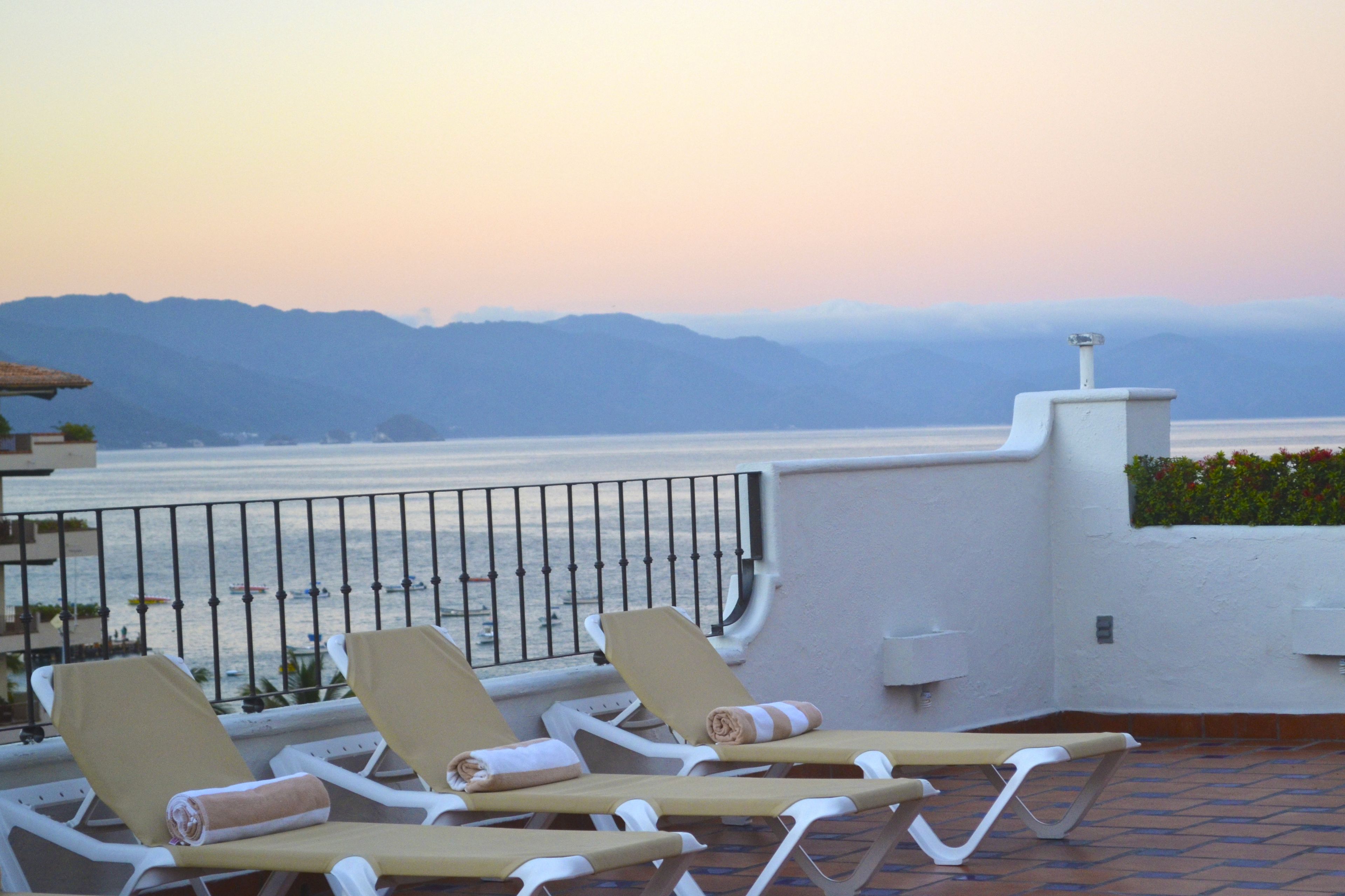 A terrace with lounge chairs and a view of the sea and mountains.