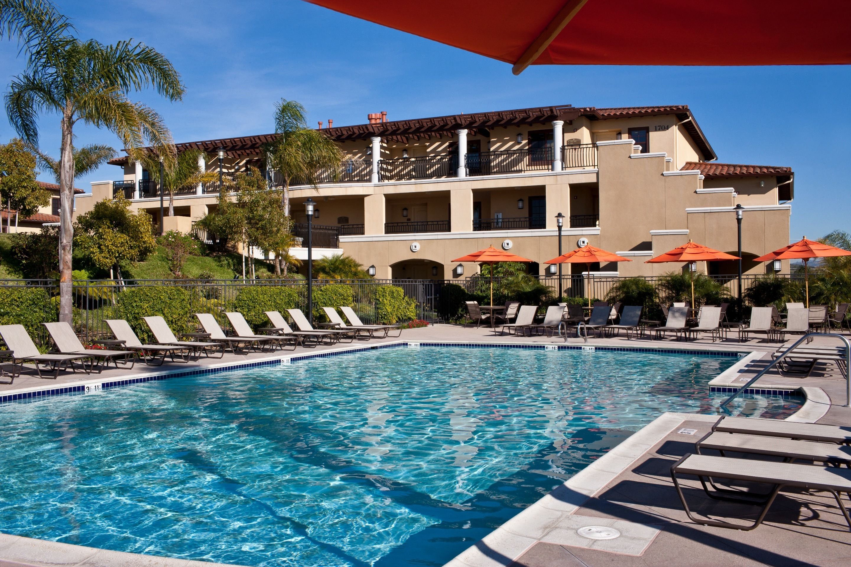 A pool area with lounge chairs and umbrellas, surrounded by a building with balconies and palm trees.