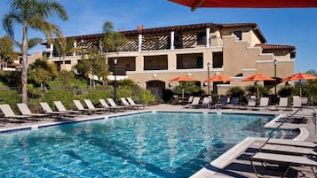 A pool area with lounge chairs and umbrellas, surrounded by a building with balconies and palm trees.