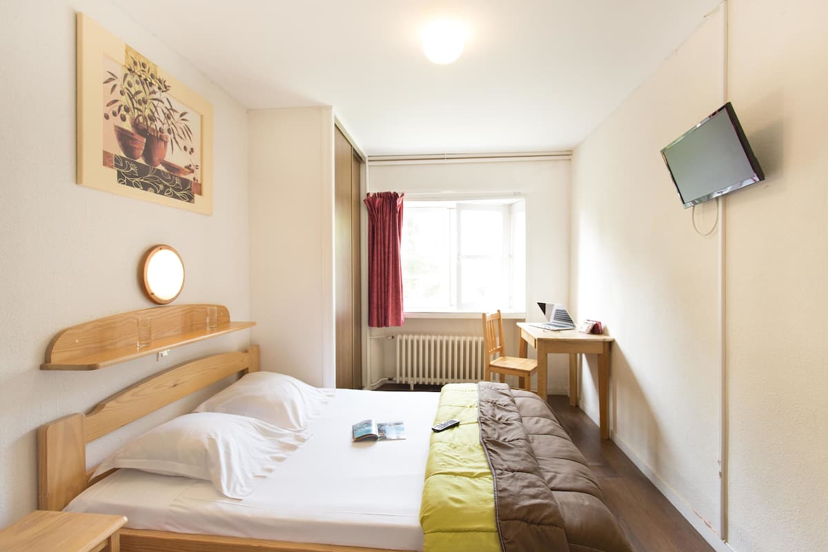 Une chambre d’hôtel avec un lit, un bureau, une chaise, une télévision, une fenêtre avec des rideaux et un tableau au mur.