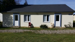 A single-story building with a dark roof, white exterior walls, and blue trim around the windows and doors. There are two doors and two windows visible, with a small garden area in front. - Ma Normandie by Popinns (Breville-les-Monts)