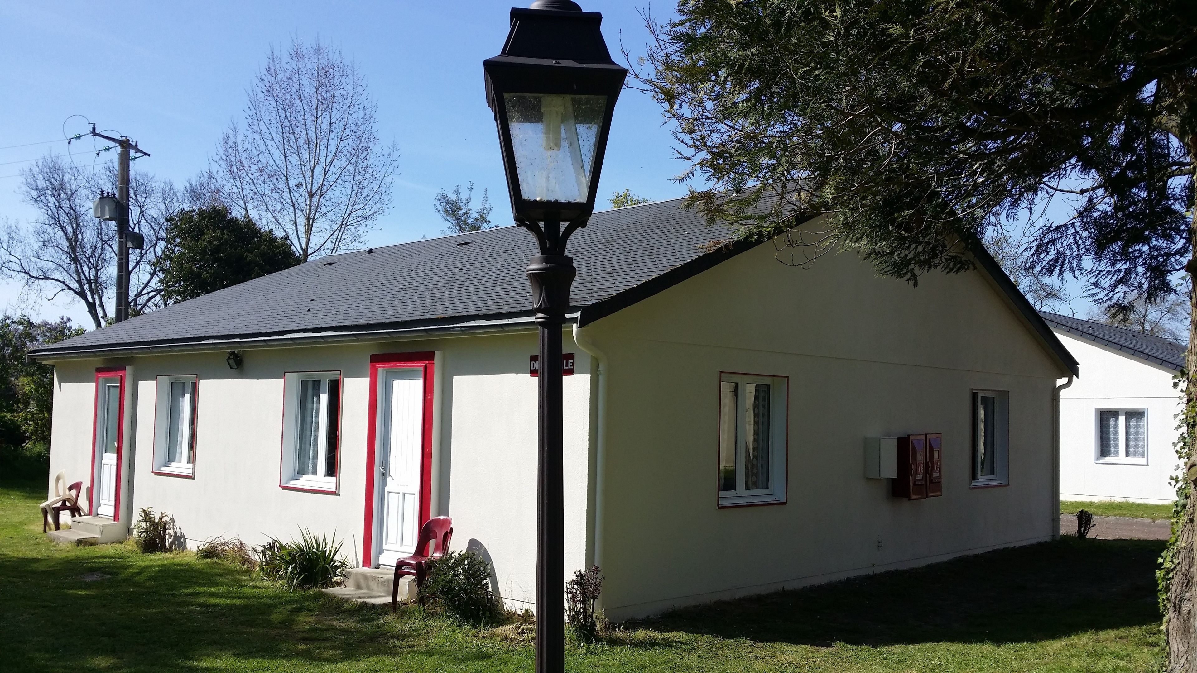 A single-story building with a sloped roof, a red door, and a small porch with a chair.