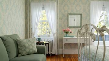 A room with a green sofa, a white bed with a headboard, a small table with a vase of flowers, and a window with white curtains.