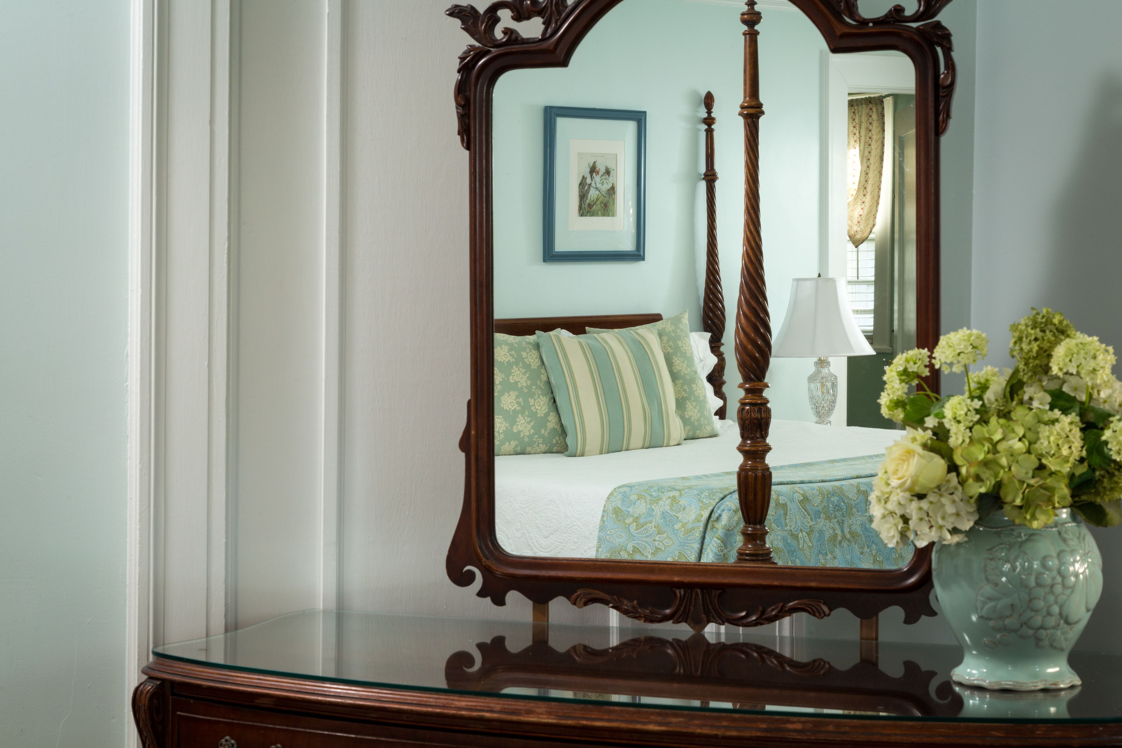 A bedroom with a four-poster bed, a vase with flowers, and a framed mirror on a dresser.
