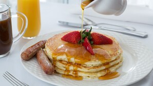 A breakfast plate with pancakes, strawberries, and syrup, accompanied by sausages and a glass of orange juice.