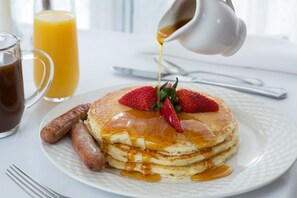 A breakfast plate with pancakes, strawberries, and syrup, accompanied by sausages and a glass of orange juice.